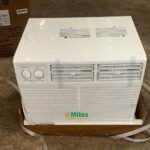 White window air conditioner with two control dials on the left and front vents mounted in a cardboard cradle on a concrete floor