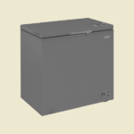 Grey compact chest freezer with a top opening lid and a thermostat dial on the lower right set against a pale beige backdrop