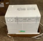 Portable white air conditioner or cooler with front vent grille and two control knobs on the left sits on a cardboard tray on a concrete floor Mitos logo visible on the front