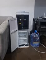 CWAY water dispenser with a small open fridge underneath placed in a living room beside a sofa with a large blue water jug nearby