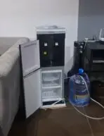 CWAY water dispenser with a small open fridge underneath placed in a living room beside a sofa with a large blue water jug nearby